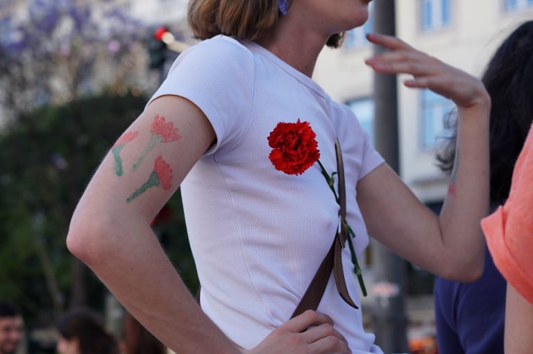 Imagem ilustrativa do evento. Fotografia com uma mulher de camisola branca com cravos vermelhos. A fotografia tem Direitos Reservados.